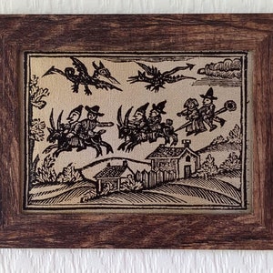 May include: A framed black and white illustration of a group of witches flying on broomsticks with a goat and a small house in the background.