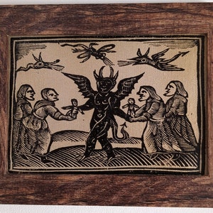 Può includere: Un'incisione su legno in bianco e nero incorniciata che raffigura un diavolo con ali e coda, che tiene una piccola bambola. Quattro figure, due donne e due bambini, interagiscono con il diavolo. L'incisione è su uno sfondo beige chiaro.