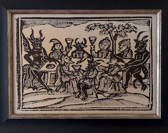 May include: A framed black and white print featuring a woodcut-style illustration of several figures, including horned creatures, gathered around a table with glasses. The artwork is enclosed in a rectangular black frame with a thin silver inner border.