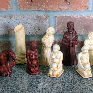 May include: A collection of nine small, hand-painted figurines. The figurines are made of a light brown material and depict various figures, including a tower, a woman, a man, and a dog. The figurines are arranged in a row on a brick surface.