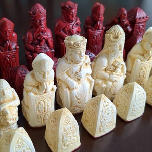 May include: A set of chess pieces featuring detailed figures in red and cream colours. The pieces include kings, queens, bishops, and knights, all designed with intricate carvings and pointed bases. The set is arranged on a dark surface.