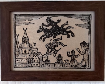 May include: A framed black and white print on textured fabric featuring a woodcut-style illustration of a horned figure riding a horse above a town, with a falling figure holding a scythe and two people standing below in a dark brown wooden frame.