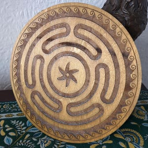 May include: A circular wooden labyrinth, featuring a star-shaped design at its core. The labyrinth is carved into the wood, with a decorative swirl pattern around the perimeter. The wood has a natural, light brown hue.