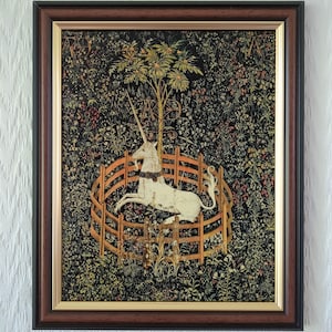 May include: A framed tapestry print featuring a white unicorn resting inside a circular wooden fence. The background is a dark, dense field of colourful wildflowers and greenery, with a central tree and the letters A and E visible in the design.