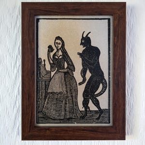 Puede incluir: Una impresión en blanco y negro enmarcada que representa a una mujer y una figura diabólica. La mujer viste un vestido largo y sostiene cartas. El diablo tiene cuernos y una cola. El marco es de madera oscura.