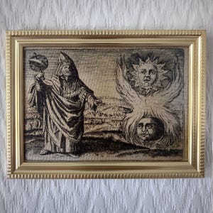 May include: A framed black and white etching featuring two distinct images. One depicts a robed figure holding an orb, the other a sun-like face with wings above a human face. The frame is gold-colored.