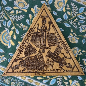 May include: A wooden, triangular plaque with intricate carvings and text. The design features a central figure and additional figures at the bottom, surrounded by detailed inscriptions. The plaque is set against a patterned green fabric.