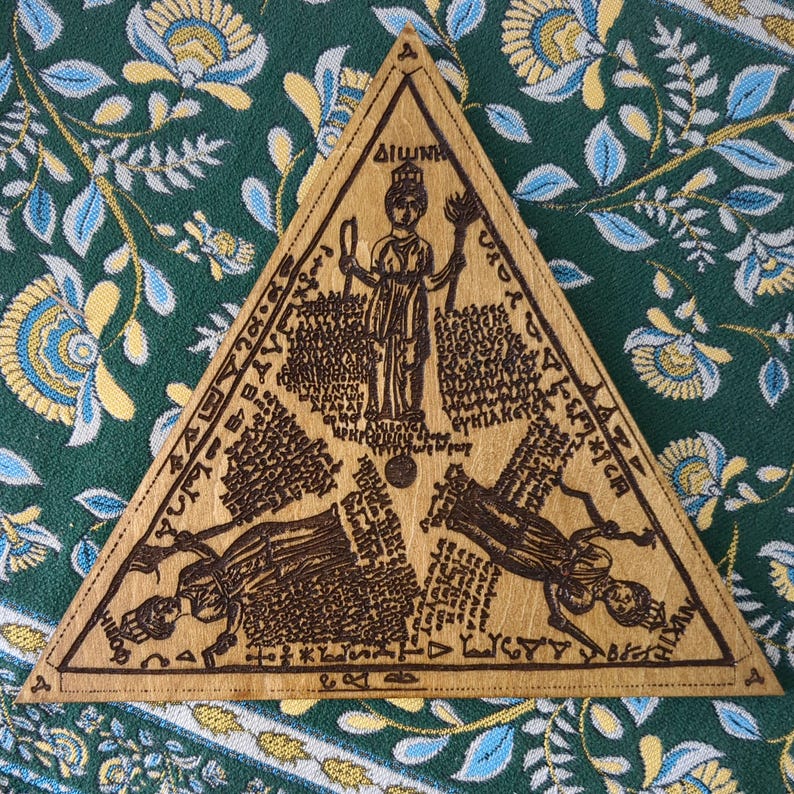 May include: A wooden, triangular plaque with intricate carvings and text. The design features a central figure and additional figures at the bottom, surrounded by detailed inscriptions. The plaque is set against a patterned green fabric.