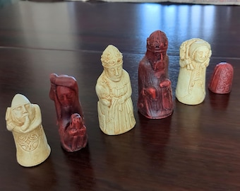 May include: A collection of intricately carved chess pieces in ivory and reddish-brown hues. The pieces depict figures with detailed facial features and clothing, showcasing a historical or fantasy theme. The set includes a bishop, queen, and other chess figures.