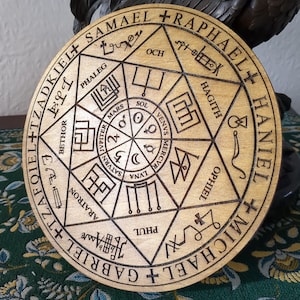 Puede incluir: Un círculo de madera con un complejo diseño geométrico grabado en él. El diseño incluye un círculo central con los nombres de los planetas y símbolos, rodeado de un círculo más grande con los nombres de los ángeles y símbolos.