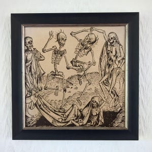 May include: A framed square wall art piece featuring a black ink illustration on a beige background. The artwork depicts four skeletons in various poses, with one playing a flute, two dancing, and one reclining on the ground, all enclosed in a black frame.