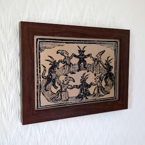 May include: A framed print featuring a black and white illustration of figures with horns and tails dancing in a circle. The artwork is set against a tan background and enclosed in a dark brown wooden frame.