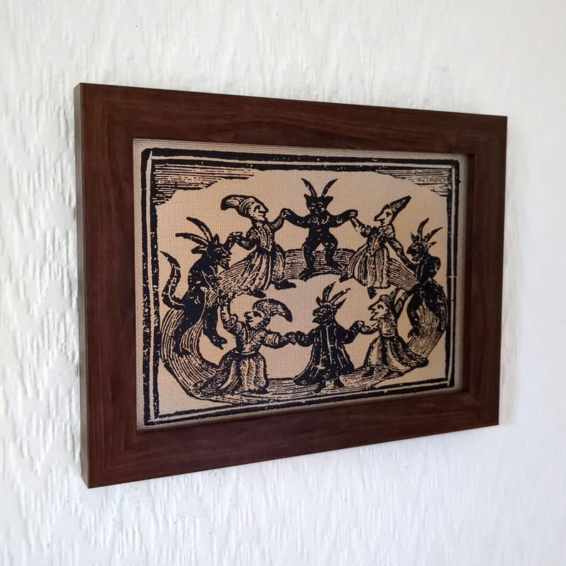 May include: A framed print featuring a black and white illustration of figures with horns and tails dancing in a circle. The artwork is set against a tan background and enclosed in a dark brown wooden frame.