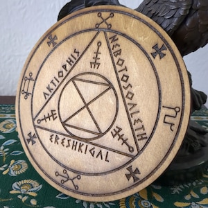 Puede incluir: Placa redonda de madera clara. El diseño intrincado incluye formas geométricas y símbolos, como un triángulo, un círculo y cruces. El texto circular dice "AKTIOPHIS", "NEROTOSOALETH" y "ERESHKIGAL".