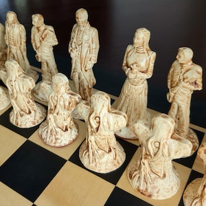 May include: A set of off-white and brown stained chess pieces arranged on a checkered board. The pieces include detailed human figures in period clothing and abstract, tentacled forms with textured surfaces, all standing on circular bases.