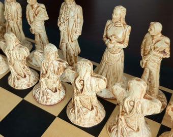 May include: A set of off-white and brown stained chess pieces arranged on a checkered board. The pieces include detailed human figures in period clothing and abstract, tentacled forms with textured surfaces, all standing on circular bases.