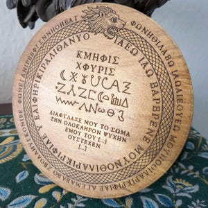 May include: A round, wooden coaster with engraved text and symbols. The coaster features a lion's head and a snake design around the edge. The coaster has Greek text and symbols in the center. The wood has a natural, light brown color.