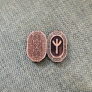 May include: Two wooden rune stones with detailed patterns. One has a complex knotwork design, the other a rune symbol within an oval frame. Both are light brown with darker etched details, set against a textured green background.