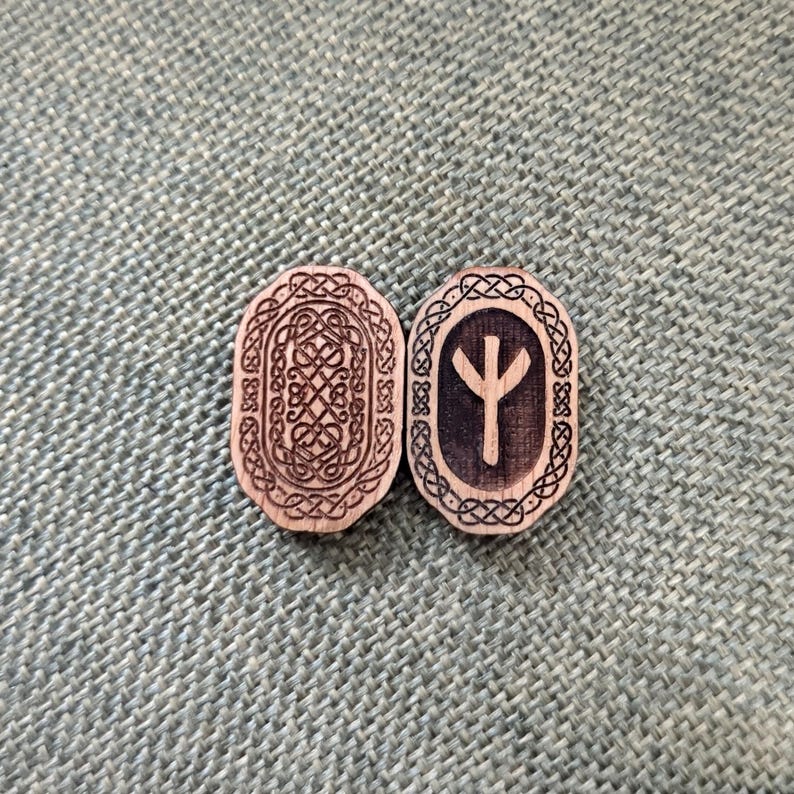 May include: Two wooden rune stones with detailed patterns. One has a complex knotwork design, the other a rune symbol within an oval frame. Both are light brown with darker etched details, set against a textured green background.