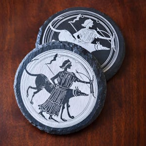 Può includere: Due sottobicchieri rotondi in ardesia nera con un disegno bianco inciso. Il disegno presenta una figura con arco e freccia, accompagnata da un cane. I sottobicchieri hanno un bordo naturale e ruvido.