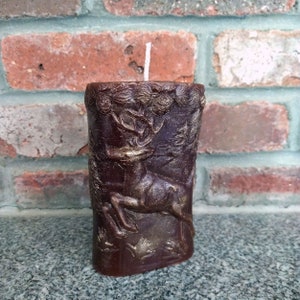 May include: A brown pillar candle with a deer design and gold accents. The candle has a white wick.