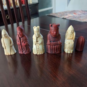 May include: A set of chess pieces in cream and reddish-brown. The pieces include a king, queen, bishop, and knight, with detailed carvings. A dark brown chess piece is also present. The pieces are arranged on a dark wooden surface.