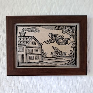 Könnte beinhalten: Ein gerahmter Schwarz-Weiß-Druck auf strukturiertem Stoff mit einer Illustration im Holzschnitt-Stil, die eine Figur zeigt, die auf ein Haus mit rauchendem Schornstein zufliegt. Das Bild befindet sich in einem dunkelbraunen Holzrahmen.