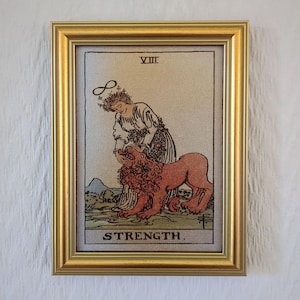 May include: A framed tarot card print featuring the Strength card. The image depicts a woman with a floral crown and a lion. The card is framed in a gold-colored frame. The card has the Roman numeral VIII at the top and the word "STRENGTH" at the bottom.