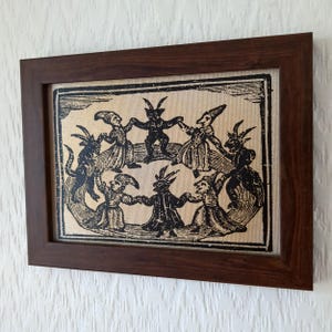 May include: A framed print featuring a black and white illustration of figures with horns and pointed hats dancing in a circle. The artwork is set against a tan background and enclosed in a dark brown wooden frame.