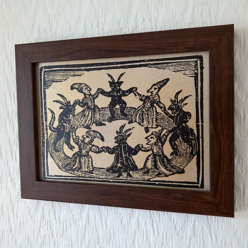May include: A framed print featuring a black and white illustration of figures with horns and pointed hats dancing in a circle. The artwork is set against a tan background and enclosed in a dark brown wooden frame.