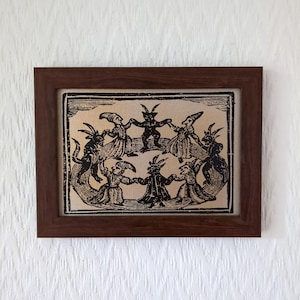 May include: A framed print featuring a vintage illustration of figures with horns and pointed hats dancing in a circle. The artwork is in black and white, set against a beige background, and framed in a dark brown wooden frame.