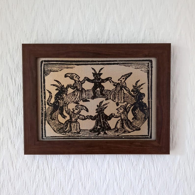 May include: A framed print featuring a vintage illustration of figures with horns and pointed hats dancing in a circle. The artwork is in black and white, set against a beige background, and framed in a dark brown wooden frame.
