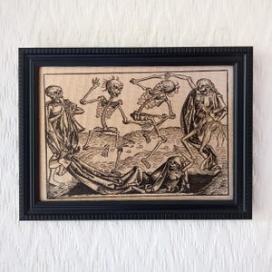 May include: A framed black and white print on a textured beige background featuring a group of five skeletons in various poses, with some wearing draped fabric, set within a simple black rectangular frame with a ridged border design.