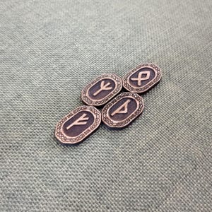 May include: Four oval-shaped, dark grey rune stones with etched symbols and decorative borders. The runes are in a light grey colour. The stones are arranged on a textured, light green surface.