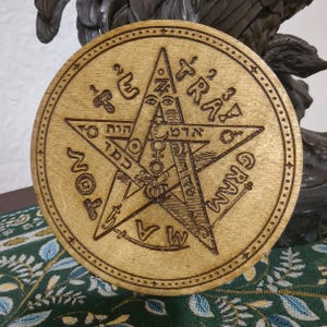 May include: A round, wooden coaster with a pentagram design. The star is etched with symbols and text, surrounded by a dotted border. The coaster is made of light-colored wood.