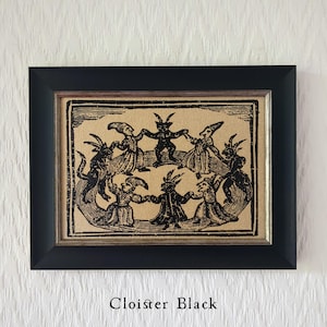 May include: Black framed artwork featuring a vintage illustration of figures with horns and cloaks dancing in a circle. The artwork is printed on a tan fabric and the text "Cloister Black" is at the bottom.
