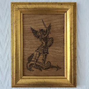 May include: A framed woodcut depicting a figure with wings, a sword, and a shield, standing over a dragon. The artwork is set against a light wood grain background and framed in a gold-colored frame.
