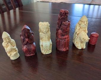 May include: A collection of intricately carved chess pieces in ivory and reddish-brown hues. The set includes figures of a king, queen, and other traditional pieces, each with unique details. The pieces are arranged on a dark wooden surface.