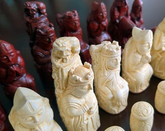 Op de afbeelding: Een verzameling schaakstukken met gedetailleerde figuren in crème en donkerrode kleuren. De set bevat uitgesneden koningen, koninginnen en lopers met fijne gelaatstrekken en kledingtexturen, naast cilindervormige stukken met spiraalvormige patronen.