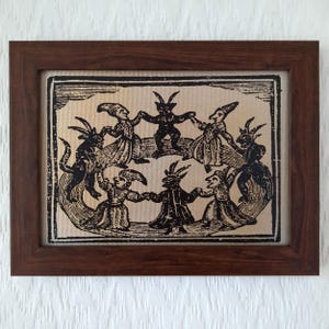May include: A framed print featuring a black and white illustration of figures dancing in a circle. The figures have horns and are dressed in robes. The frame is made of dark brown wood, and the print is on a beige background.
