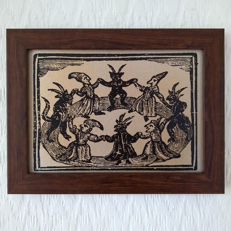 May include: A framed print featuring a black and white illustration of figures dancing in a circle. The figures have horns and are dressed in robes. The frame is made of dark brown wood, and the print is on a beige background.