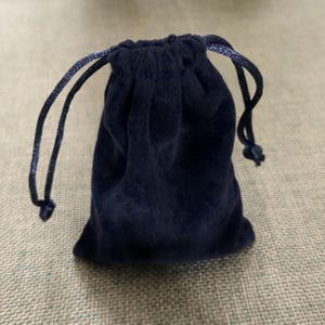 May include: A navy blue velvet drawstring pouch. The pouch is closed with a matching blue cord. The bag is small and appears to be made of a soft material. The background is a light tan.