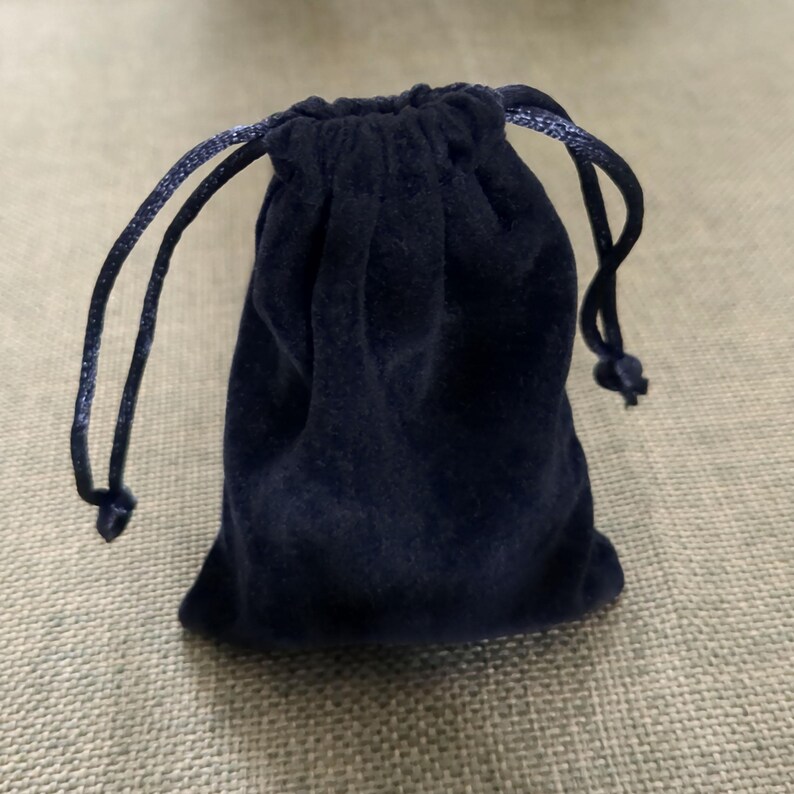 May include: A navy blue velvet drawstring pouch. The pouch is closed with a matching blue cord. The bag is small and appears to be made of a soft material. The background is a light tan.