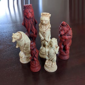 Può includere: Un set di pezzi degli scacchi scolpiti con animali, nei colori crema e rosso. Include un leone, un orso, un gatto e una scimmia. I pezzi sono esposti su una superficie marrone scuro, evidenziando i dettagli.