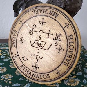 May include: A round wooden plaque with a brown stain. The plaque has a circle inscribed with the words "MICHAEL SADAY ATHANATOS" and "SABAOth". Inside the circle is a complex symbol with the letters "S" and "M" in the center.
