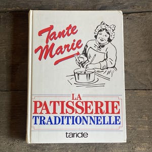 Op de afbeelding: Een wit hardcover boek met de titel Tante Marie La Patisserie Traditionnelle en de uitgeversnaam Taride onderaan. De kaft bevat een zwart-witte lijntekening van een persoon die in een pan roert en rode en blauwe tekst.