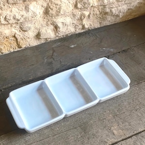 May include: A white, rectangular serving dish with three sections. The dish has rounded edges and small handles. It is made of ceramic or porcelain and is on a wooden surface. The dish is ideal for serving snacks.