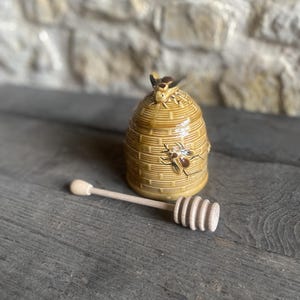 Könnte beinhalten: Beigefarbener Keramik-Honigtopf in Bienenkorbform, verziert mit Bienenfiguren. Ein Honiglöffel aus Holz liegt neben dem Topf. Der Hintergrund ist eine Steinmauer. Der Löffel und der Topf sind hellbraun.