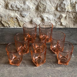 May include: A collection of eight vintage pink-tinted drinking glasses. The glasses are cylindrical with a faceted base, arranged on a weathered wooden surface. The background features a textured stone wall.
