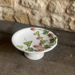 May include: A white ceramic cake stand with a raised base. The plate is decorated with green leaves and pink strawberries. The stand is on a wooden surface.